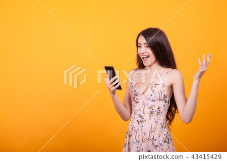 Happy exited pretty girl in short dress holding mobile phone in studio over yellow background Happy exited pretty girl in short dress holding mobile phone in studio over yellow background 43415429