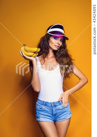 Woman in swimsuit and blue shorts holding banana and posing isolated over yellow background 43424893