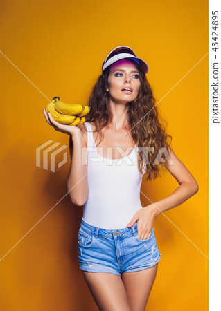 Woman in swimsuit and blue shorts holding banana and posing isolated over yellow background 43424895