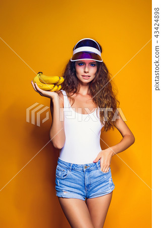 Woman in swimsuit and blue shorts holding banana and posing isolated over yellow background 43424898