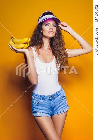 Woman in swimsuit and blue shorts holding banana and posing isolated over yellow background 43424902