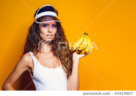Woman in swimsuit and blue shorts holding banana and posing isolated over yellow background 43424907