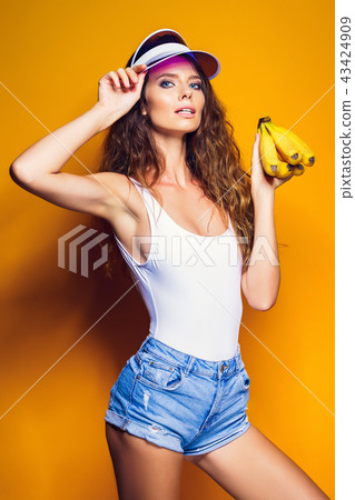 Woman in swimsuit and blue shorts holding banana and posing isolated over yellow background 43424909