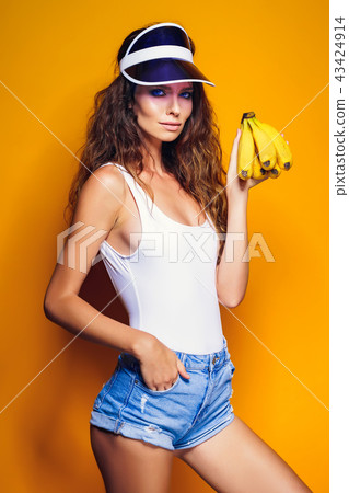 Woman in swimsuit and blue shorts holding banana and posing isolated over yellow background 43424914