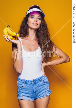 Woman in swimsuit and blue shorts holding banana and posing isolated over yellow background 43424922