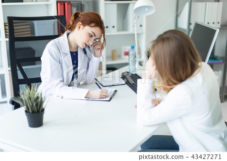 A beautiful young girl in a white robe is sitting at the desk in the office and communicating with 43427271