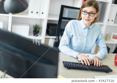 A young girl in glasses sits at a table in the office, holds a pencil in her hand and types the text 43427316