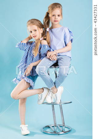 Girls twins in light blue clothes are posing near a bar stool on a blue background. Girls twins in light blue clothes are posing near a bar stool on a blue background. 43427331