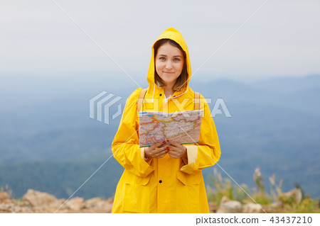 Hipster young girl in yellow raincoat with backpack on peak of mountain, looking map. Tourist Hipster young girl in yellow raincoat with backpack on peak of mountain, looking map. Tourist 43437210