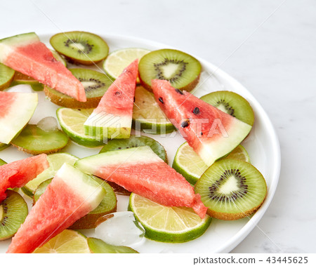 Pattern of pieces of watermelon, kiwi, lime and melting ice cubes in a plate on a gray marble 43445625