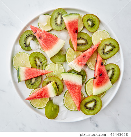 Juicy slices of fruit, lime, kiwi, watermelon and ice cubes in a plate on a gray marble background Juicy slices of fruit, lime, kiwi, watermelon and ice cubes in a plate on a gray marble background 43445638