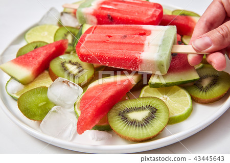 Close-up woman's hand holds berry ice cream lolly, on a white background a plate of ice cream 43445643