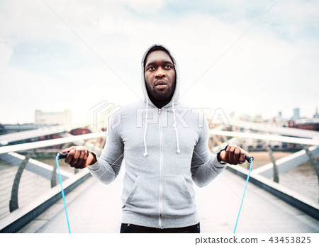 Young active black sportsman skipping with a rope in a city, wearing hoodie. 43453825