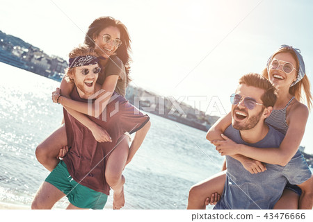 Outdoors photo of happy boyfriends piggybacking their girlfriends at sunset on beach 43476666