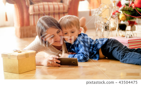 Portrait of little boy watching cartoons on mobile phone with mother under Christmas tree 43504475