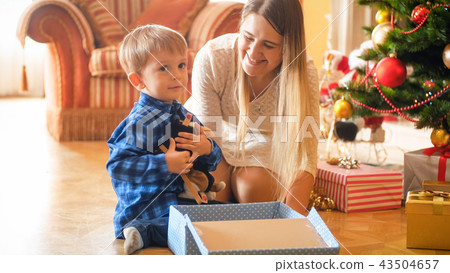 Adorable little boy sitting under Christmas tree and hugging plush toy he received in gift box from Adorable little boy sitting under Christmas tree and hugging plush toy he received in gift box from 43504657