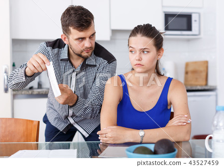 Upset young man with bill having conflict with girl in home interior Upset young man with bill having conflict with girl in home interior 43527459