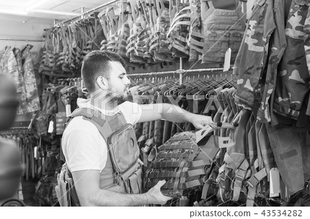 Young guy choosing flak jacket in military shop Young guy choosing flak jacket in military shop 43534282