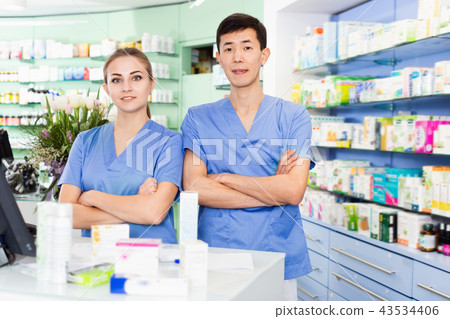 Portrait of two pharmacists who are standing on their work place near table with cashbox in apothecary. 43534406