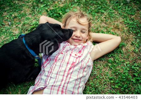 A small girl lying down on grass, a black dog licking her face. 43546403