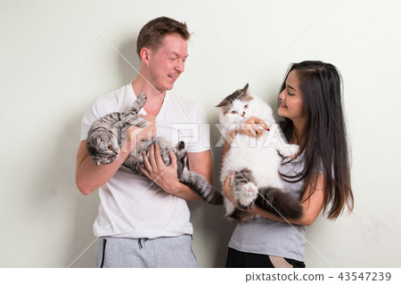 Young happy multi-ethnic couple holding two cats together against white background Young happy multi-ethnic couple holding two cats together against white background 43547239