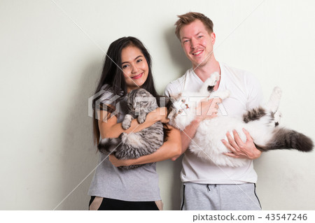 Young happy multi-ethnic couple holding two cats together against white background Young happy multi-ethnic couple holding two cats together against white background 43547246
