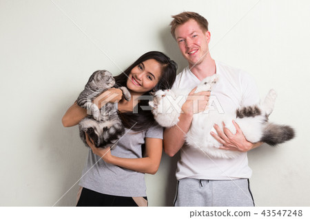 Young happy multi-ethnic couple holding two cats together against white background Young happy multi-ethnic couple holding two cats together against white background 43547248