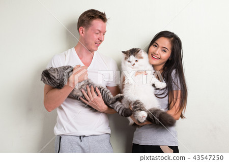 Young happy multi-ethnic couple holding two cats together against white background Young happy multi-ethnic couple holding two cats together against white background 43547250