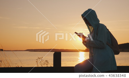 A woman in a hood sits on a fence near a picturesque lake, uses a smartphone. Beautiful sunset A woman in a hood sits on a fence near a picturesque lake, uses a smartphone. Beautiful sunset 43638348