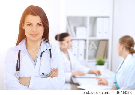 Brunette female doctor on the background of colleagues talking to each other in hospital. Medicine Brunette female doctor on the background of colleagues talking to each other in hospital. Medicine 43646038