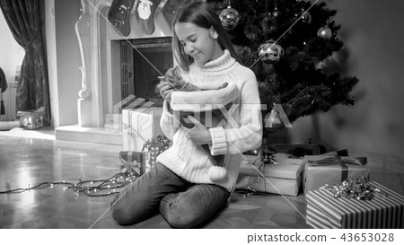 Black and white photo of beautiful smiling girl in sweater sitting with cat next to decorated Black and white photo of beautiful smiling girl in sweater sitting with cat next to decorated 43653028