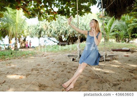 Young girl riding swing and making selfie by smartphone, sand and trees in background. 43663603