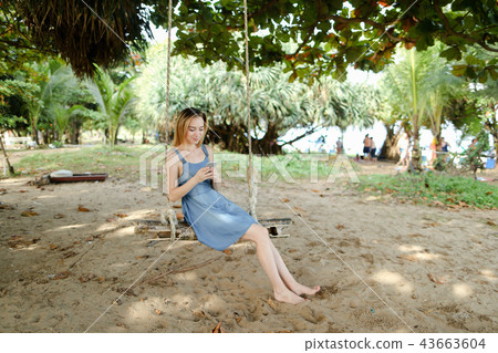 Young caucasian woman riding swing and using smatrphone, sand and tree in background. 43663604