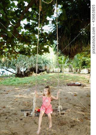 Little funny girl riding on swing on sand background, wearing pink dress. 43663605