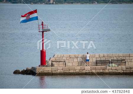 Croatian flag flying in wind on lighthouse in harbor, daytime, stone pier, man, facing away looking Croatian flag flying in wind on lighthouse in harbor, daytime, stone pier, man, facing away looking 43782848