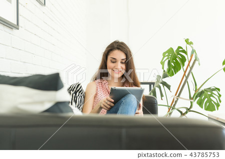 Technologies, communication, people concept - young woman is sitting on sofa and chatting in tablet Technologies, communication, people concept - young woman is sitting on sofa and chatting in tablet 43785753