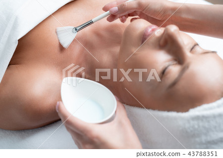 Beautician using cosmetic brush for applying mask on woman skin Beautician using cosmetic brush for applying mask on woman skin 43788351