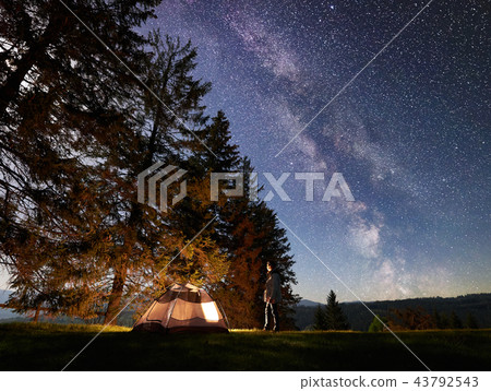 Male hiker enjoyng night camping near tourist tent at campfire under blue starry sky and Milky way Male hiker enjoyng night camping near tourist tent at campfire under blue starry sky and Milky way 43792543