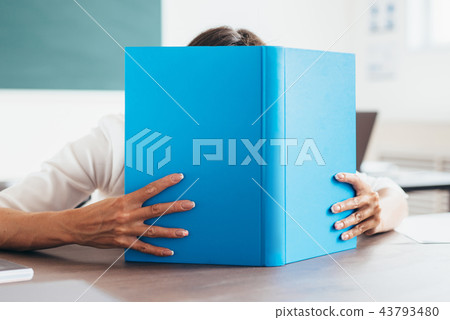 Student girl covering her face behind a book in classroom. 43793480