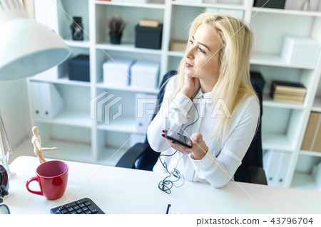 A young girl in the office sits at a desk holding a phone in her hand and talking on it through a A young girl in the office sits at a desk holding a phone in her hand and talking on it through a 43796704