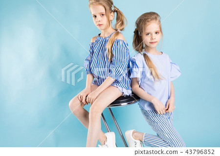 Girls twins in light blue clothes are posing near a bar stool on a blue background. Girls twins in light blue clothes are posing near a bar stool on a blue background. 43796852