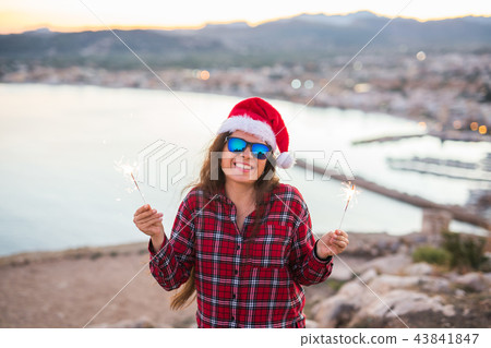 Christmas, holidays, people concept - woman in christmas hat and glasses holding sparklers over sea Christmas, holidays, people concept - woman in christmas hat and glasses holding sparklers over sea 43841847