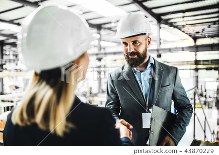 A portrait of an industrial man and woman engineer in a factory, shaking hands. A portrait of an industrial man and woman engineer in a factory, shaking hands. 43870229