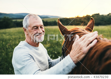 A senior man standing close to a horse outdoors in nature, stroking it. 43870230