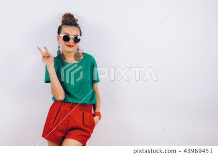 beautiful woman in sunglasses wearing in red shorts and green T-shirt standing near white wall beautiful woman in sunglasses wearing in red shorts and green T-shirt standing near white wall 43969451