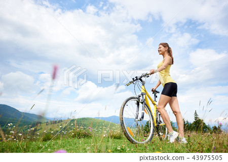 Attractive female cyclist with yellow mountain bicycle, enjoying sunny day in the mountains Attractive female cyclist with yellow mountain bicycle, enjoying sunny day in the mountains 43975505