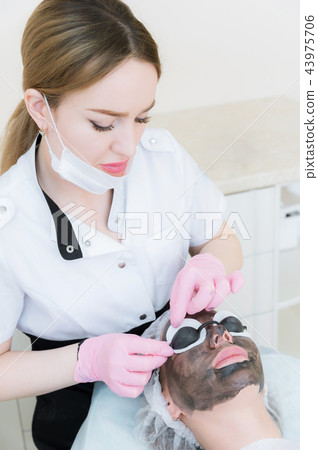 The cosmetologist in pink gloves with a brush applies a carbon mask for peeling on the face of a The cosmetologist in pink gloves with a brush applies a carbon mask for peeling on the face of a 43975706
