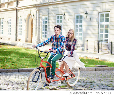 Young tourist couple, handsome man and pretty blond woman riding tandem bicycle along city street. 43976475