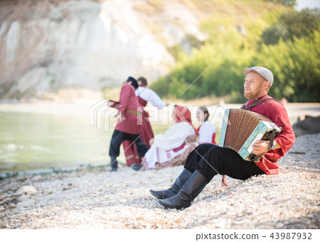 In the foreground, a man in a Russian folk costume sits and plays the accordion against a beautiful In the foreground, a man in a Russian folk costume sits and plays the accordion against a beautiful 43987932