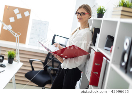 A young girl stands near the shelf and holds a folder with documents in her hands. A young girl stands near the shelf and holds a folder with documents in her hands. 43996635